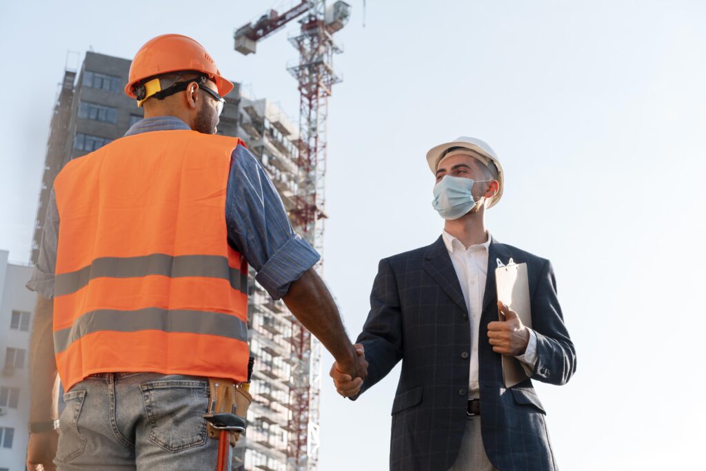 Construction worker and engineer shaking hands at a construction site wearing safety helmets.